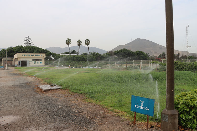 Sistema de riego por aspersión funcionando en campo de cultivo de la UNALM, con edificio de Caseta de Riego al fondo